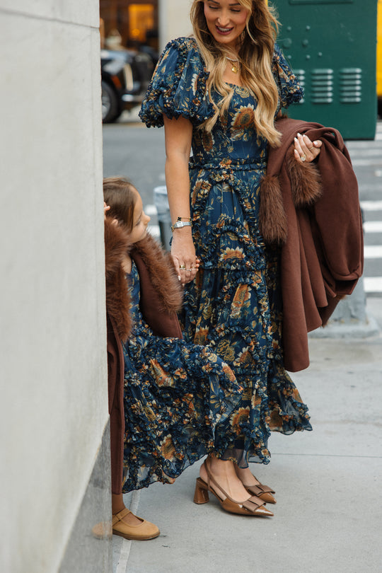 A woman and a young girl, both wearing the Ivy City Co Mini Marseille Dress in Sunflower Twilight—a matching blue floral dress ideal as a girls midi dress or for holiday portraits—with brown shoes, stand hand-in-hand on a city sidewalk. Each carries a brown coat with fur trim draped over her arm.