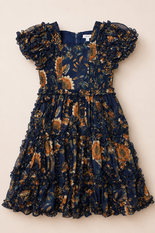 The Mini Marseille Dress in Sunflower Twilight by Ivy City Co is a navy blue floral dress featuring yellow flowers, short puff sleeves, a gathered bodice, ruffled trim, and a tiered skirt. Laid flat on a beige background, this charming piece makes the perfect girls holiday dress.