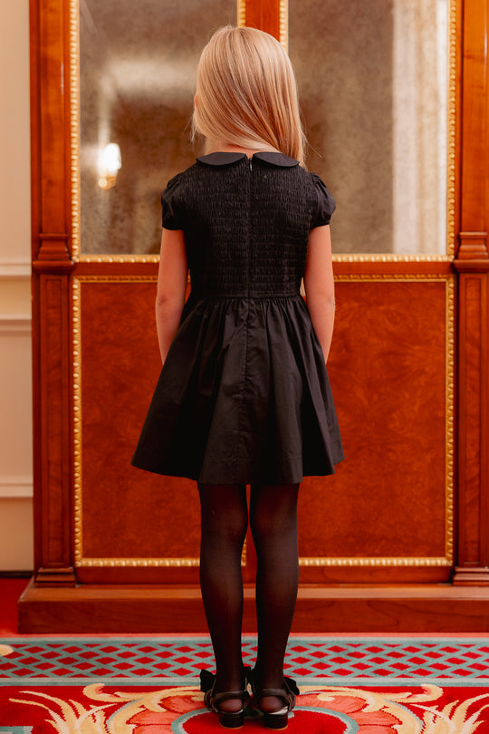 the back of a little girl in a black dress standing on a colorful carpet. 