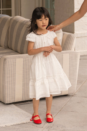 A young girl wears the Ivy City Co Mini Palermo Dress in White with puff sleeves and red shoes, standing indoors near a striped couch as an adult hand reaches toward her face.