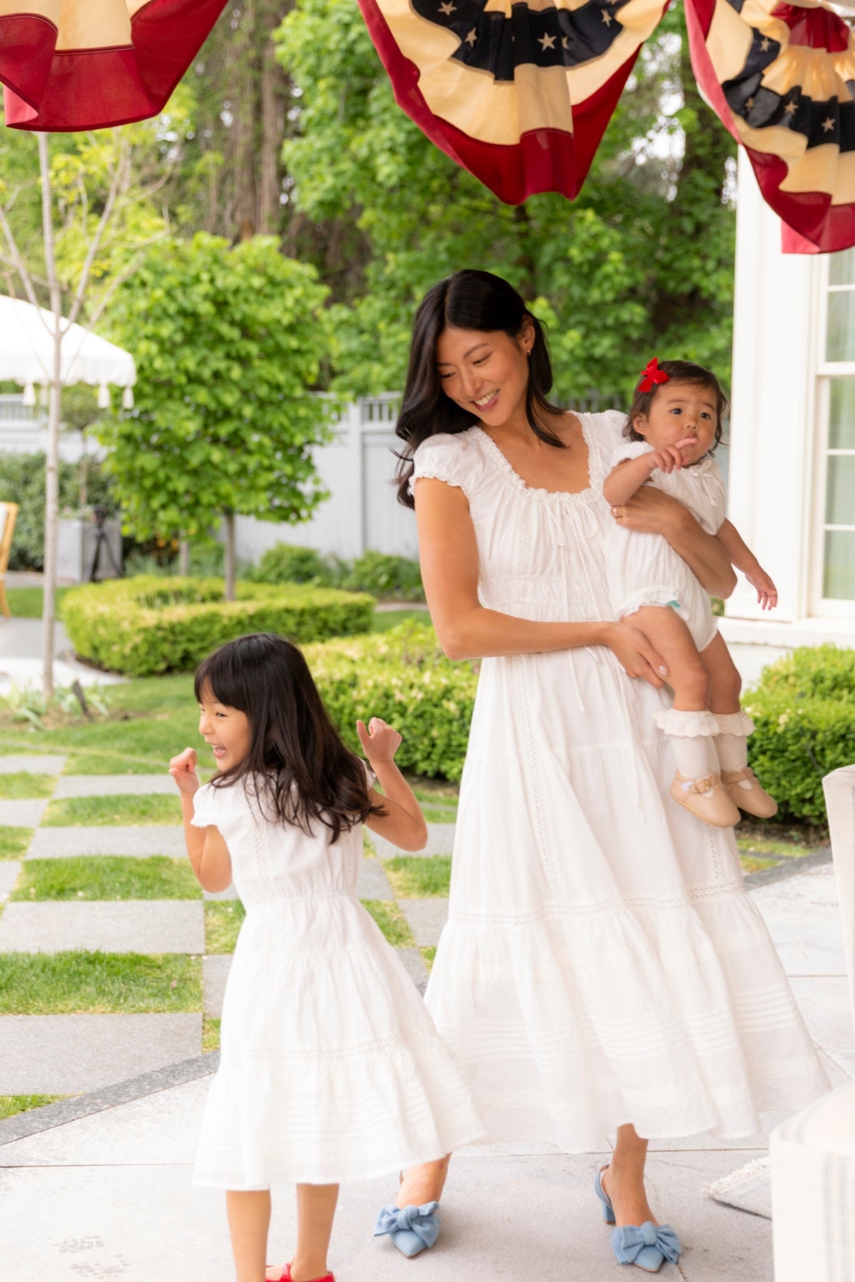 A woman in the Ivy City Co Mini Palermo Dress in White smiles while holding a baby and watching a girl twirl. They are outdoors on a patio decorated with greenery and red, white, and blue patriotic bunting overhead.