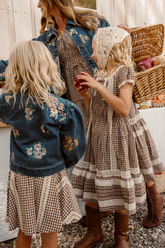 Three girls in brown gingham dresses, one wearing a floral denim jacket and another a white crochet bonnet, stand outdoors by a table with a basket of apples. One of the girls is dressed in the Mini Palermo Dress in Hickory Gingham from Ivy City Co, featuring sweet puff sleeves and crafted from a soft cotton-blend fabric.