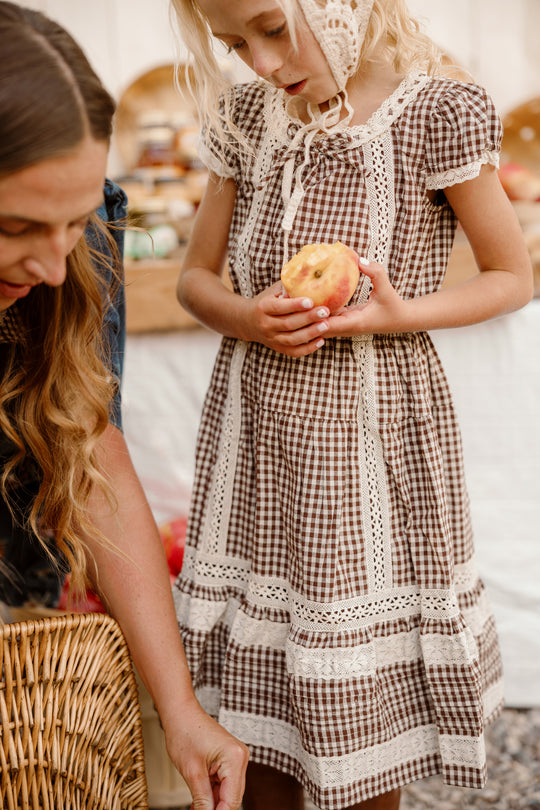 A young girl in the Ivy City Co Mini Palermo Dress in Hickory Gingham, featuring puff sleeves and delicate lace details, holds a bitten apple while standing next to a woman with long hair. Behind her, a wicker basket and a food-laden table enhance the charm of the scene, highlighting her cotton-blend outfit.