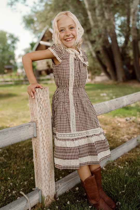 A smiling young girl wearing the Ivy City Co Mini Palermo Dress in Hickory Gingham, paired with a lace bonnet and brown boots, leans on a wooden fence in a grassy outdoor setting with trees and a house in the background.