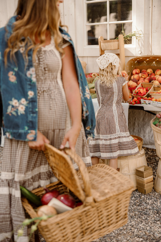 A woman holding a wicker basket filled with vegetables stands beside a girl in the Ivy City Co Mini Palermo Dress in Hickory Gingham with puff sleeves and a headscarf, who is reaching for peaches at an outdoor market.