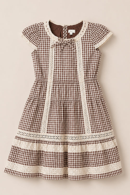 The Ivy City Co Mini Palermo Dress in Hickory Gingham features a brown and white gingham pattern, short puff sleeves, delicate lace trim, a round neckline with a small bow accent, and a tiered skirt crafted from a soft cotton-blend. The dress is shown laid flat on a neutral background.