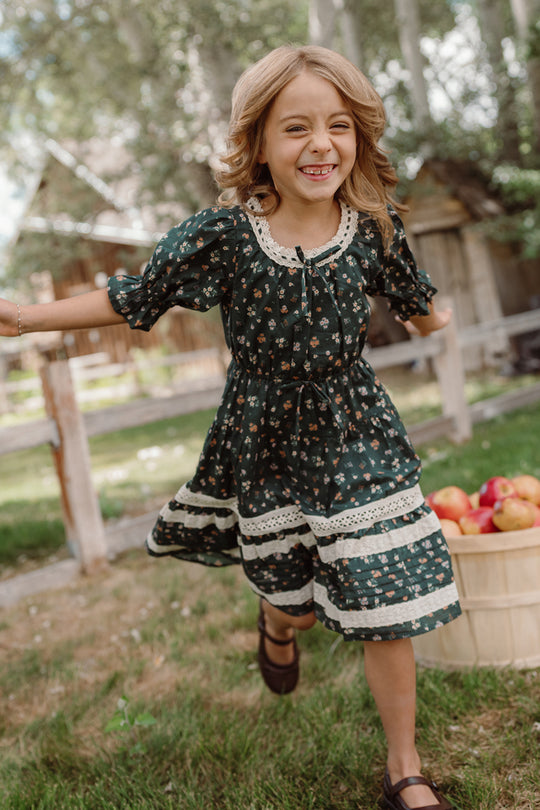A young girl beams with joy as she runs outdoors near a wooden basket filled with apples, wearing the Ivy City Co Mini Palermo 3/4 Sleeves Dress in Evergreen Lovelorn Ditsy. Trees and rustic wooden buildings provide a charming backdrop.