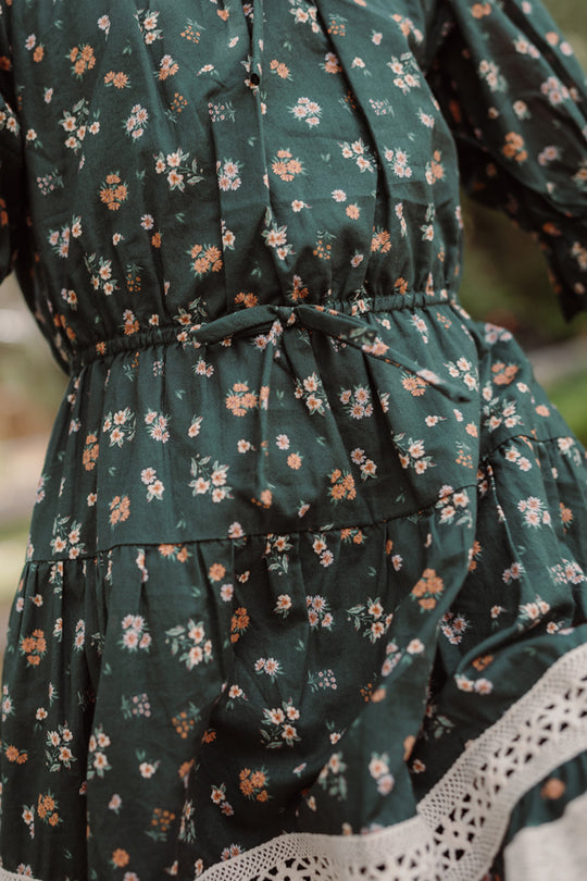 Close-up of the Ivy City Co Mini Palermo 3/4 Sleeves Dress in Evergreen Lovelorn Ditsy, showcasing its dark green shade with a delicate floral print, gathered waist, and white lace trim at the hem. This girls’ cotton dress features a slightly wrinkled texture for extra charm.