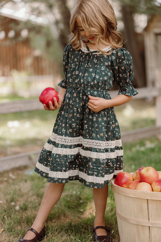 A young girl in the Ivy City Co Mini Palermo 3/4 Sleeves Dress in Evergreen Lovelorn Ditsy holds a red apple and looks down, standing beside a basket filled with apples outdoors on a sunny day.
