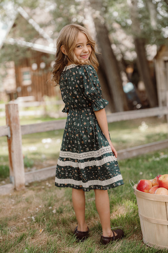 A young girl wearing the Ivy City Co Mini Palermo 3/4 Sleeves Dress in Evergreen Lovelorn Ditsy smiles over her shoulder while standing outside near a wooden fence and a basket of apples. Trees and a rustic wooden building are visible in the background.