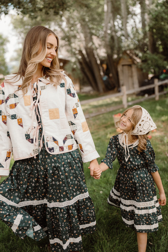 A woman and a young girl, both wearing matching Ivy City Co Mini Palermo 3/4 Sleeves Dresses in Evergreen Lovelorn Ditsy, smile at each other outdoors on green grass near trees and a wooden fence as they hold hands.
