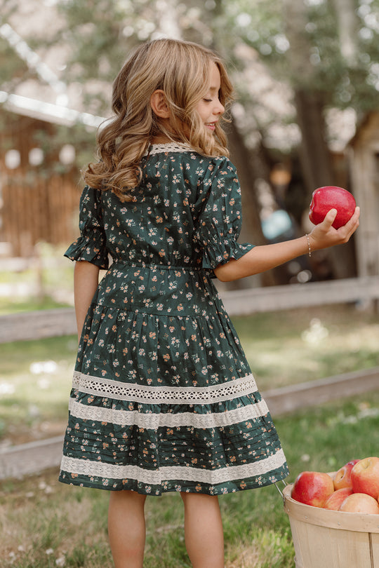 A young girl wearing the Ivy City Co Mini Palermo 3/4 Sleeves Dress in Evergreen Lovelorn Ditsy stands outdoors, holding a red apple and facing away. Nearby sits a basket brimming with apples. The grassy setting features trees and a wooden fence in the background.