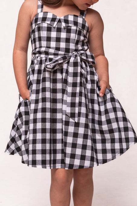 A young girl models the Ivy City Co Mini Patty Dress in Black Gingham—a sleeveless, knee-length style with a waist bow and pockets—standing against a plain background.