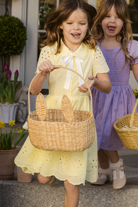 Two young girls smile and run outdoors, one wearing the Ivy City Co Mini Penny Dress in Butter Yellow Eyelet with a bunny-ear wicker basket, the other in a purple eyelet dress holding a plain basket. Flowers and greenery brighten the background.