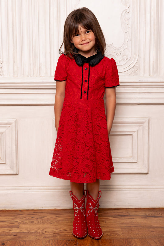 Young girl wearing a red dress with black collar and red boots standing against a white wall.