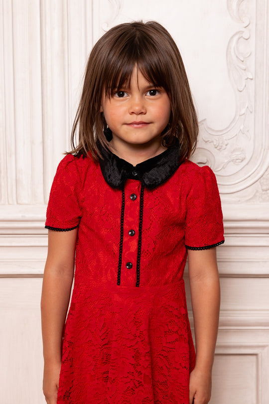 Young girl wearing a red dress with black collar and buttons against a white wall.