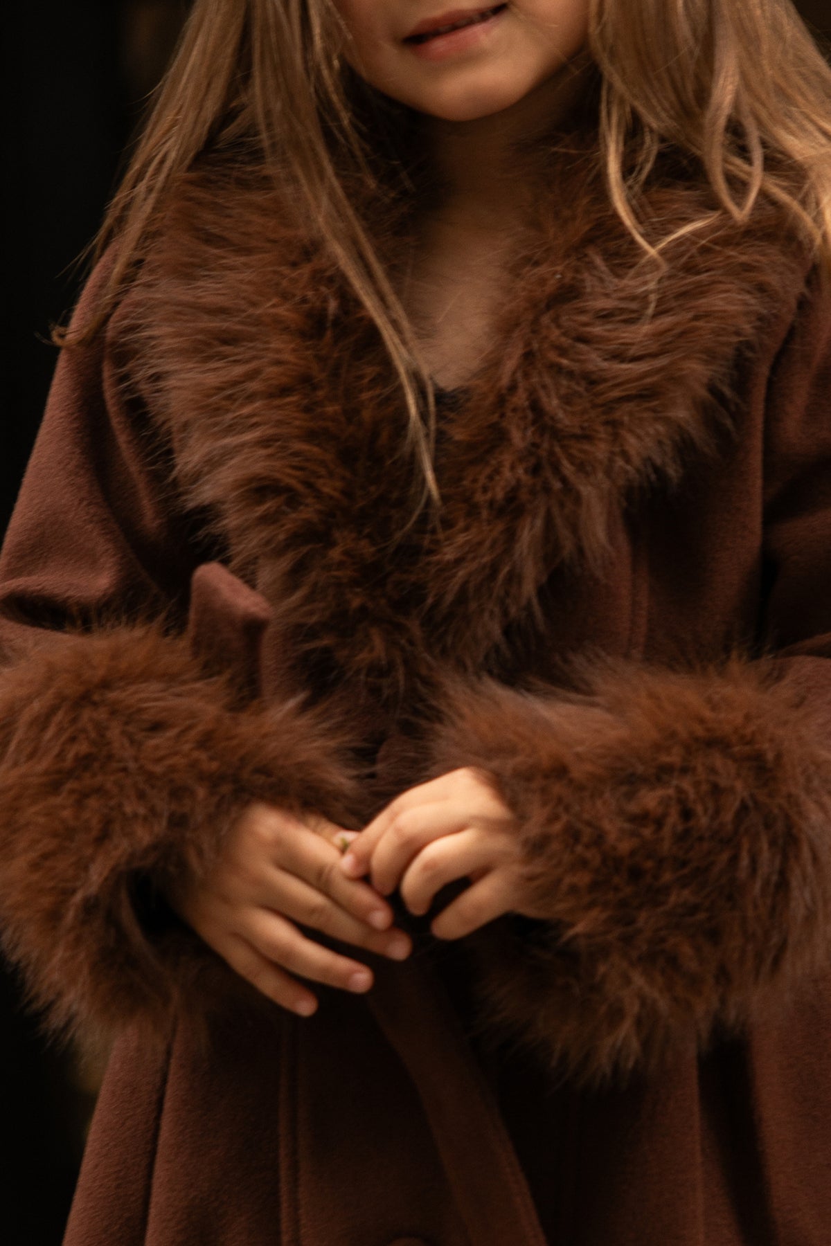 A young girl with blonde hair wears the Ivy City Co Mini Pookie Coat in Warm Cocoa, featuring a cozy design with fluffy fur trim at the collar and cuffs, her hands clasped together.