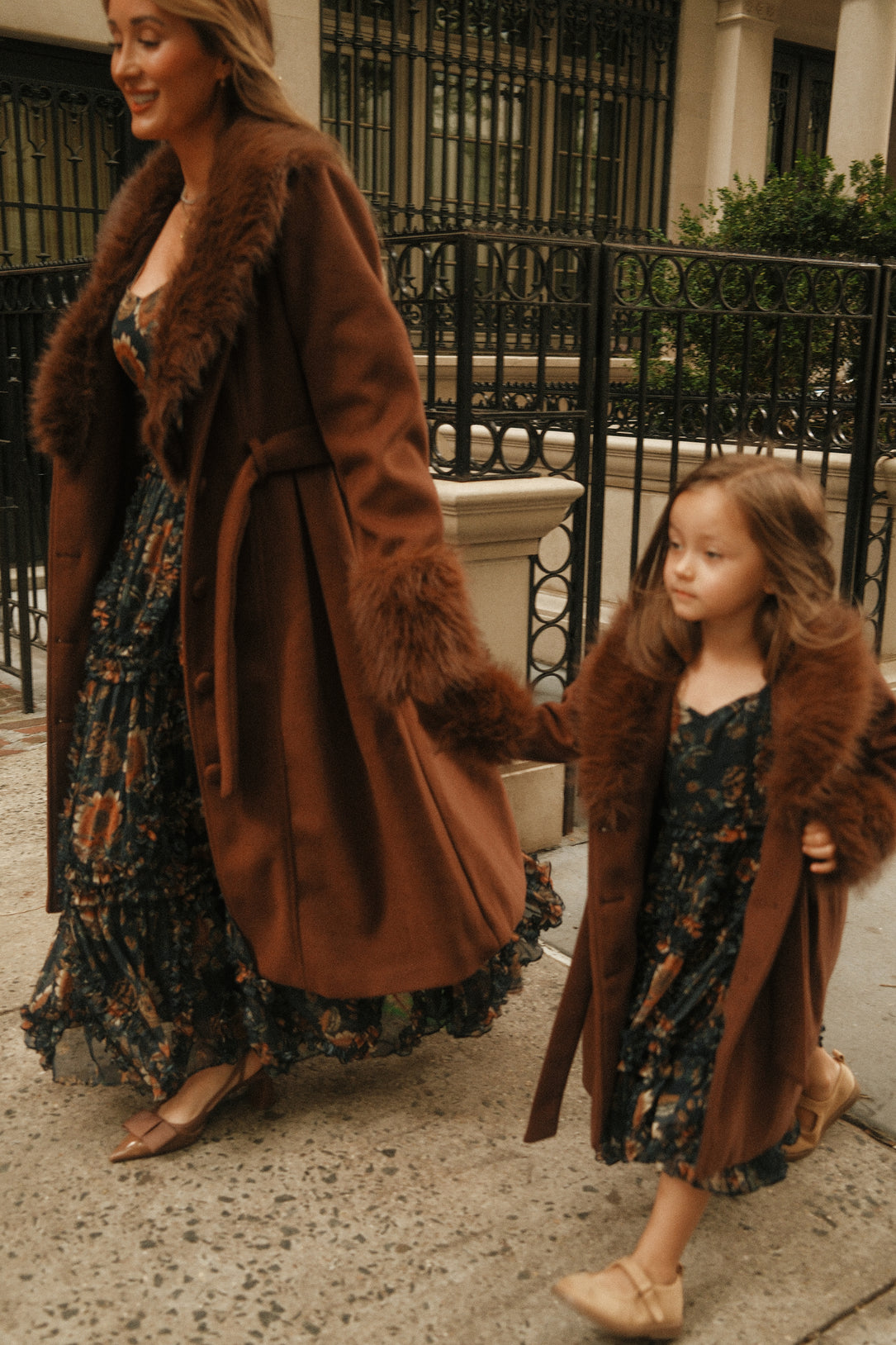 A woman and a young girl stroll hand in hand along a city sidewalk, both dressed in long, brown coats with fur trim—the young girl’s chic ensemble featuring the Ivy City Co Mini Pookie Coat in Warm Cocoa—layered over matching floral dresses.