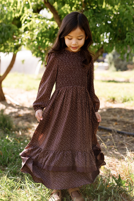 A young girl wearing the Ivy City Co Mini Presley Maxi Dress in Mahogany Ditsy stands outdoors on the grass, gently holding her skirt. The dress features a smocked bodice, elegant balloon sleeves, and a charming white ditsy print. She has long dark hair and gazes softly downward, with green trees in the background.