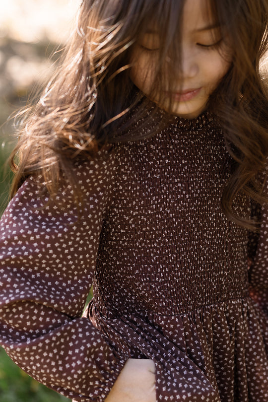 A young child with long brown hair stands outdoors, looking down with a slight smile, wearing the Ivy City Co Mini Presley Maxi Dress in Mahogany Ditsy, featuring delicate balloon sleeves and a smocked bodice.