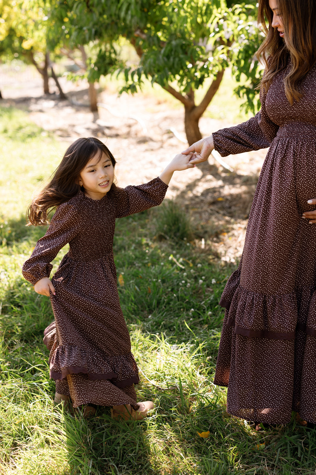 A young girl wearing the Ivy City Co Mini Presley Maxi Dress in Mahogany Ditsy, with balloon sleeves, twirls outside and holds hands with a woman dressed in a matching style featuring a smocked bodice. They are surrounded by green grass and trees on a sunny day.