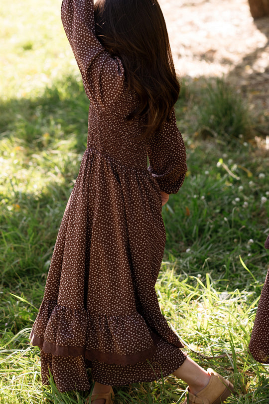 A young girl with long brown hair, wearing the Ivy City Co Mini Presley Maxi Dress in Mahogany Ditsy—featuring balloon sleeves and a smocked bodice—walks through green grass with her back to the camera on a sunny day.