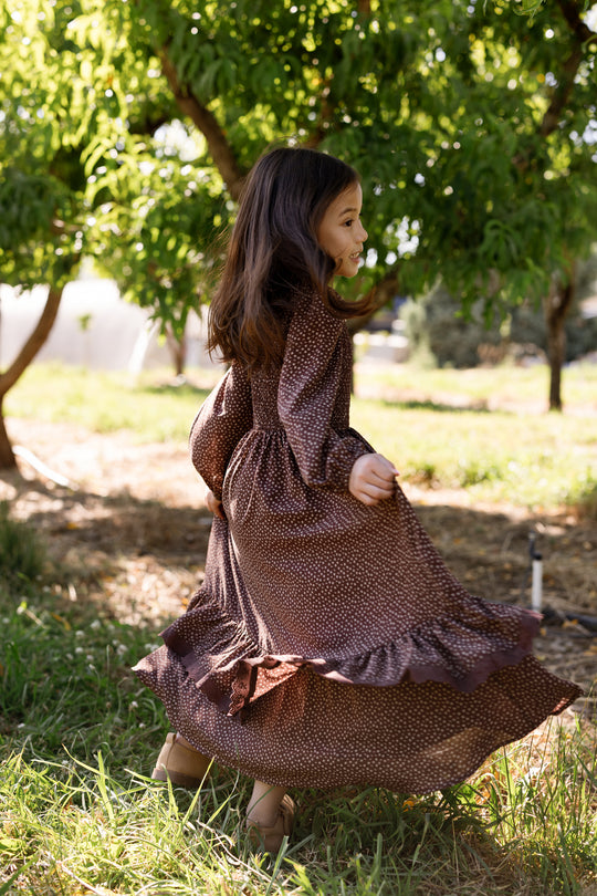 A young girl twirls joyfully outdoors near green trees, sunlight filtering through the leaves, wearing the Ivy City Co Mini Presley Maxi Dress in Mahogany Ditsy. The long, brown patterned dress features charming balloon sleeves and a smocked bodice that adds vintage elegance to her playful moment.