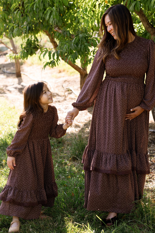 A woman and a young girl, both wearing matching Ivy City Co Mini Presley Maxi Dresses in Mahogany Ditsy with elegant balloon sleeves and a smocked bodice, hold hands and smile at each other while standing outdoors near green trees.