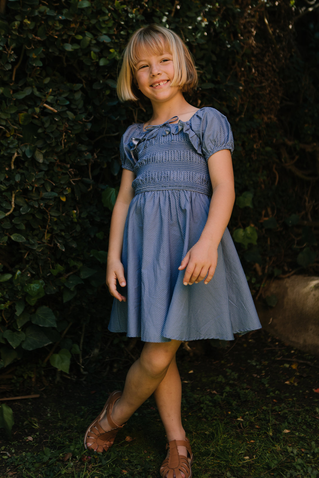 A young girl with short blonde hair smiles outdoors, wearing the Ivy City Co Mini Rae Dress in Blue Polka Dot and brown sandals, surrounded by green foliage and sunlight.