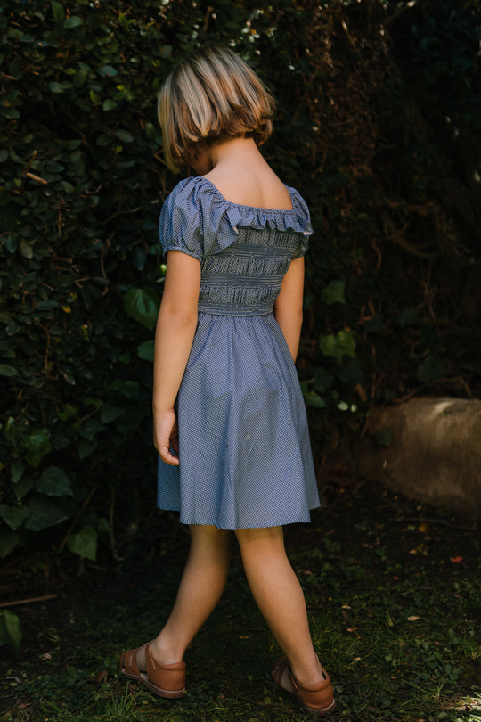 A young child with short blonde hair wears the Ivy City Co Mini Rae Dress in Blue Polka Dot and brown shoes, standing on grass near green bushes with their head turned down and back to the camera.