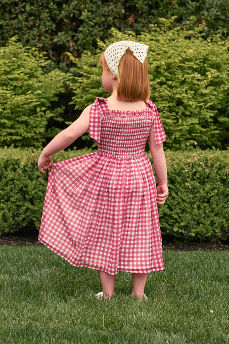 A young child with red hair and a white bow headband stands on grass, holding out one side of her Ivy City Co Mini Rayna Dress in Red Gingham. Green bushes and hedges are visible in the background.