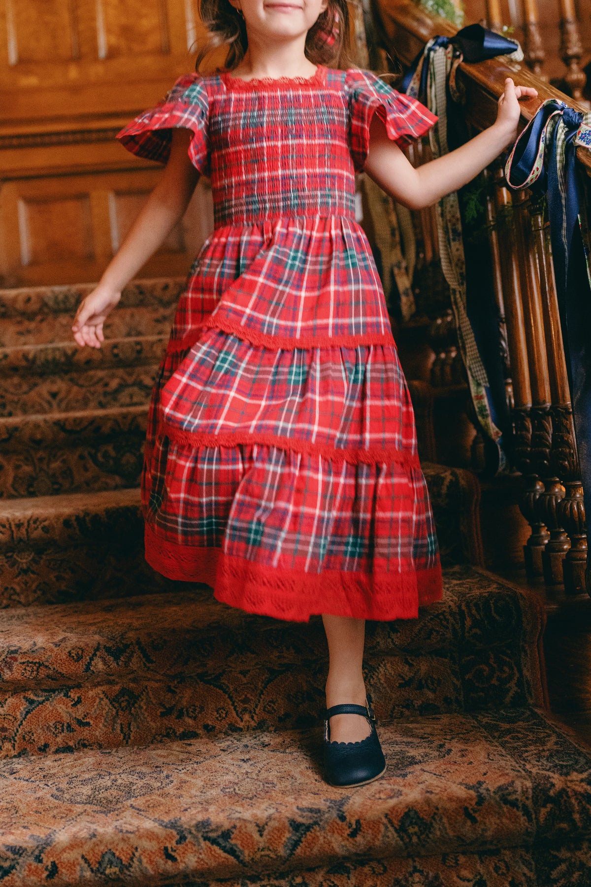 A child wearing the Ivy City Co Mini Rosario Dress in Christmas Cheer Plaid with a smocked bodice and black shoes walks down carpeted stairs, holding a wooden railing decorated with ribbon.