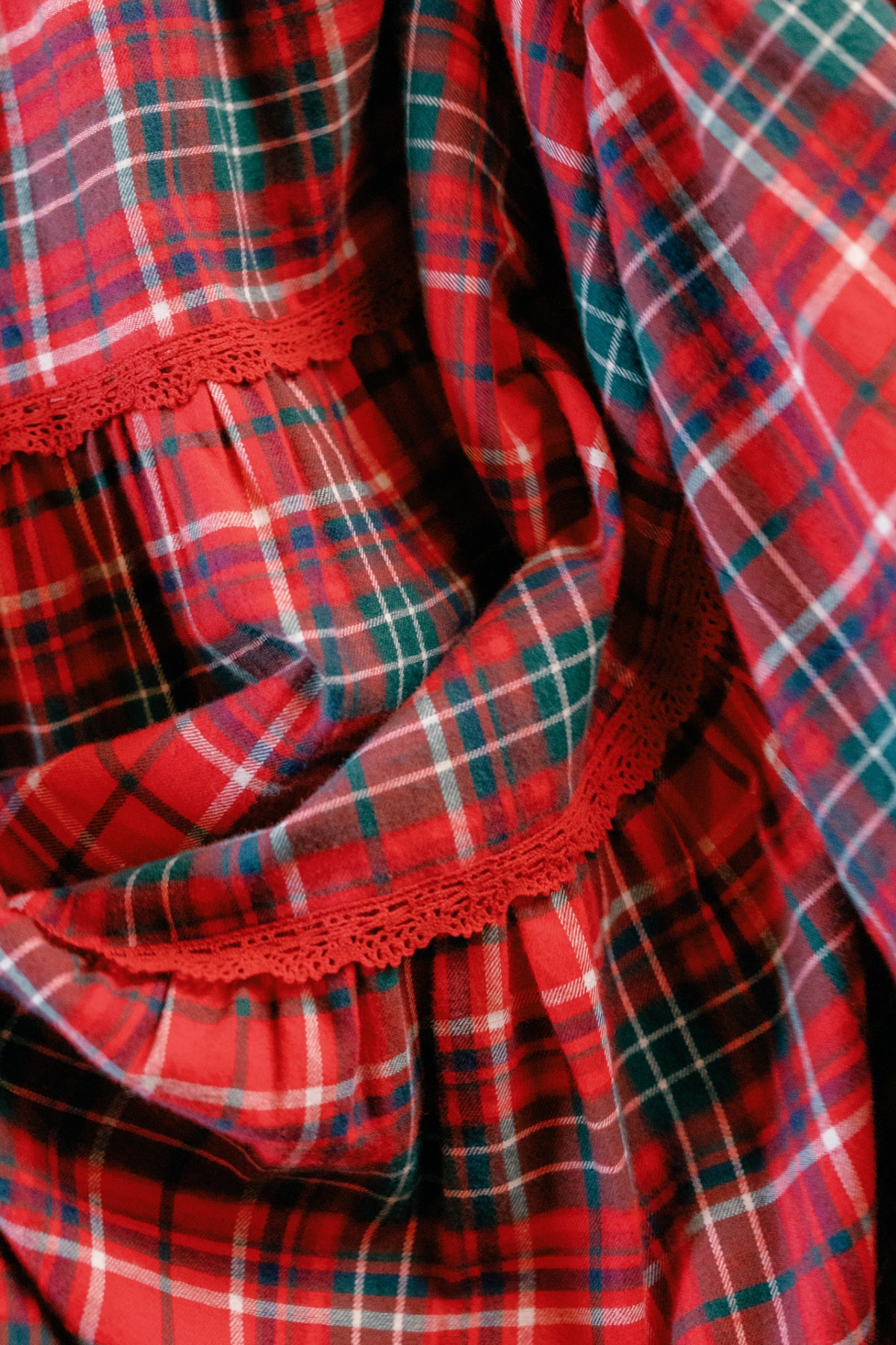 Close-up of the Ivy City Co Mini Rosario Dress in Christmas Cheer Plaid, featuring soft red, green, and white plaid fabric accented with a decorative red lace trim—showcasing delicate folds and texture that make it ideal for a festive holiday dress with a charming touch.