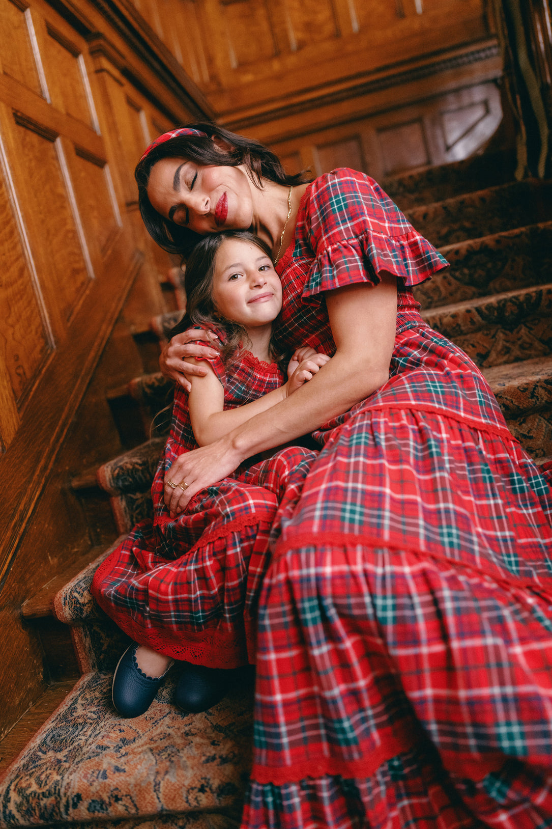 A woman and young girl sit on carpeted stairs, hugging and smiling with eyes closed, wearing matching Ivy City Co Mini Rosario Dresses in Christmas Cheer Plaid, surrounded by wood-paneled walls.