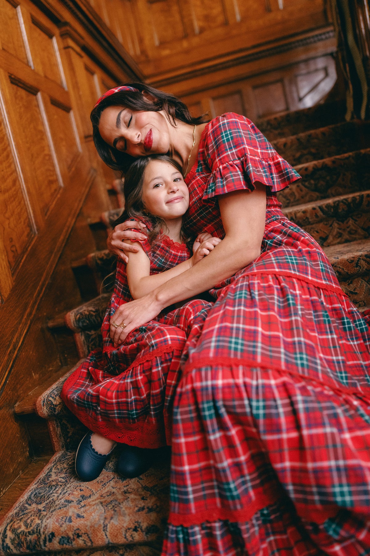 A woman and young girl sit on carpeted stairs, hugging and smiling with eyes closed, wearing matching Ivy City Co Mini Rosario Dresses in Christmas Cheer Plaid, surrounded by wood-paneled walls.