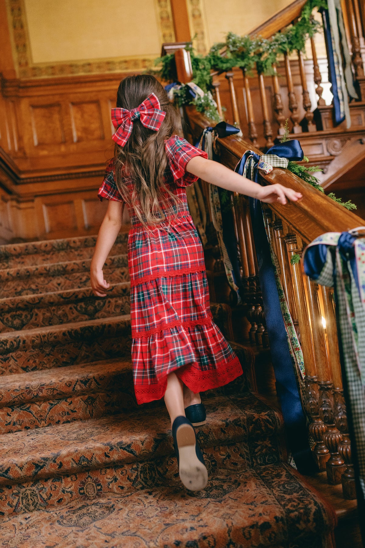A young girl wearing the Ivy City Co Mini Rosario Dress in Christmas Cheer Plaid, featuring a smocked bodice and a matching bow, climbs a carpeted staircase adorned with festive garlands.