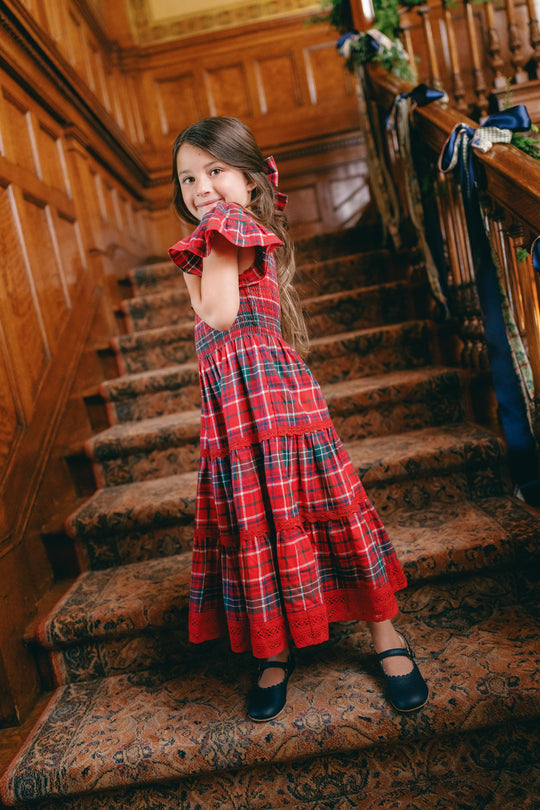 A smiling young girl with long brown hair stands on a grand wooden staircase, wearing the Ivy City Co Mini Rosario Dress in Christmas Cheer Plaid. The festive holiday dress features a smocked bodice and a red and green plaid tiered maxi skirt with ruffled sleeves. Her look is finished with a matching bow in her hair and paired with dark blue Mary Jane shoes.