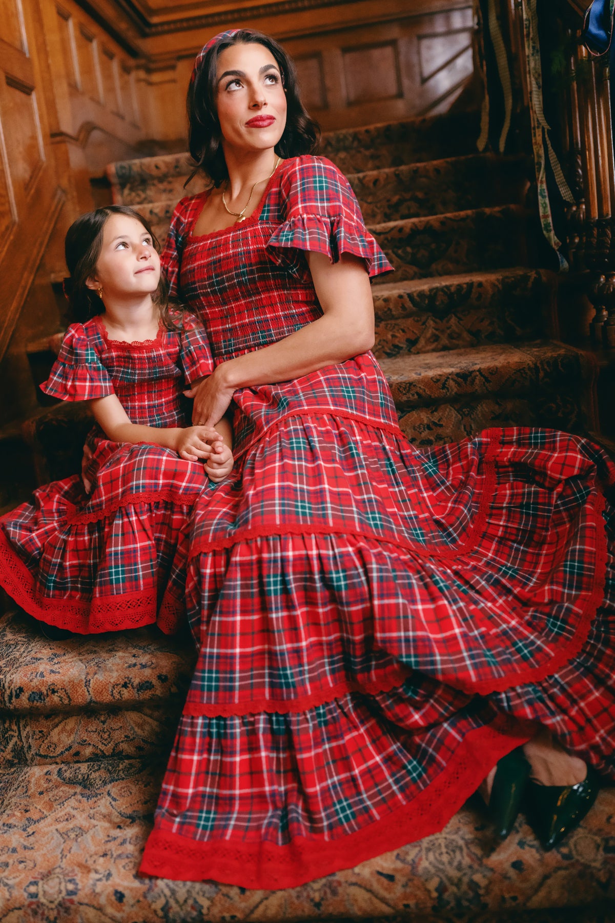 A woman and a young girl sit together on carpeted stairs, holding hands and gazing upward, each dressed in matching Ivy City Co Mini Rosario Dresses in Christmas Cheer Plaid. With their dark hair and shared smiles, they capture the heartwarming tradition of coordinated holiday style, perfectly set against wood-paneled staircase surroundings.