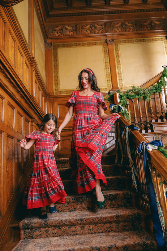 A woman and young girl, both wearing the Ivy City Co Mini Rosario Dress in Christmas Cheer Plaid with smocked bodices, walk down a staircase draped in greenery within a wood-paneled, vintage-inspired setting.