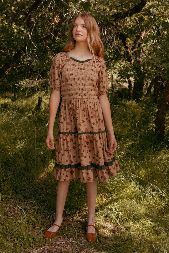 A young girl stands on grass among trees, wearing the Ivy City Co Mini Roselyn Dress in Desert Blossom—100% cotton with puffed sleeves and lace accents—paired with brown flats. She gazes slightly upward with a calm expression.