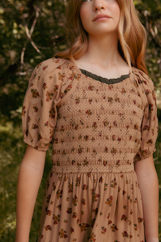 A young woman wears the Ivy City Co Mini Roselyn Dress in Desert Blossom, featuring puff sleeves and a floral pattern. Pictured outdoors with only her torso and part of her face visible, she highlights the charm of this 100% cotton dress.