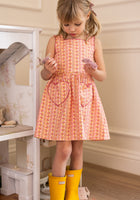 A young girl stands indoors in the Ivy City Co Mini Rue Play Dress in Handpainted Flora Stripe, holding small toys and wearing bright yellow rain boots. Her light brown hair is styled in pigtails as she looks down, ready for fun.