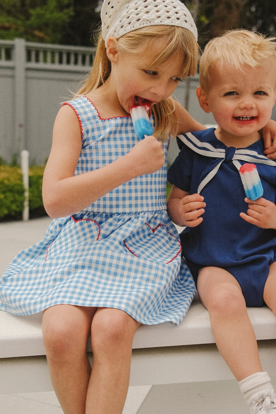 Two young children smile on a bench outdoors. The girl on the left wears the Ivy City Co Mini Rue Play Dress in Vichy Gingham and a white headscarf, enjoying a red, white, and blue popsicle beside a boy in a navy sailor outfit.
