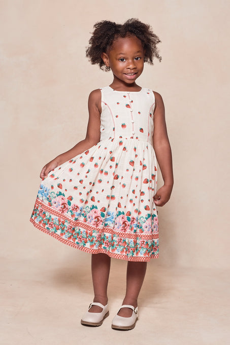 A young girl smiles in the Ivy City Co Mini Sadie Dress in Strawberry Vintage Tier—a sleeveless white cotton dress tiered with red strawberries and colorful florals at the hem. She stands on a neutral background, holding her dress and wearing white shoes.