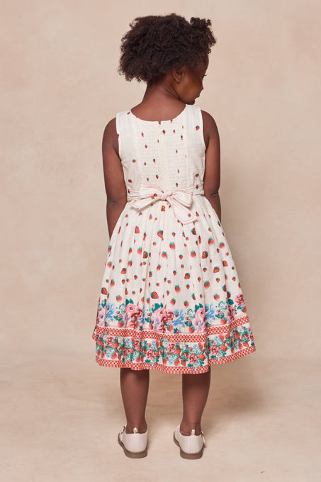 A young girl with curly hair faces away, wearing Ivy City Co's Mini Sadie Dress in Strawberry Vintage Tier, featuring red polka dots, waist bow, floral hem details, and white shoes on a beige background.