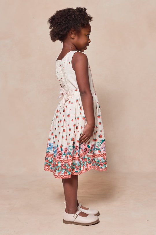 A young girl with curly hair stands sideways against a neutral background, wearing the Mini Sadie Dress in Strawberry Vintage Tier by Ivy City Co—a sleeveless, cream-colored cotton dress with tiers and colorful floral and polka dot patterns.