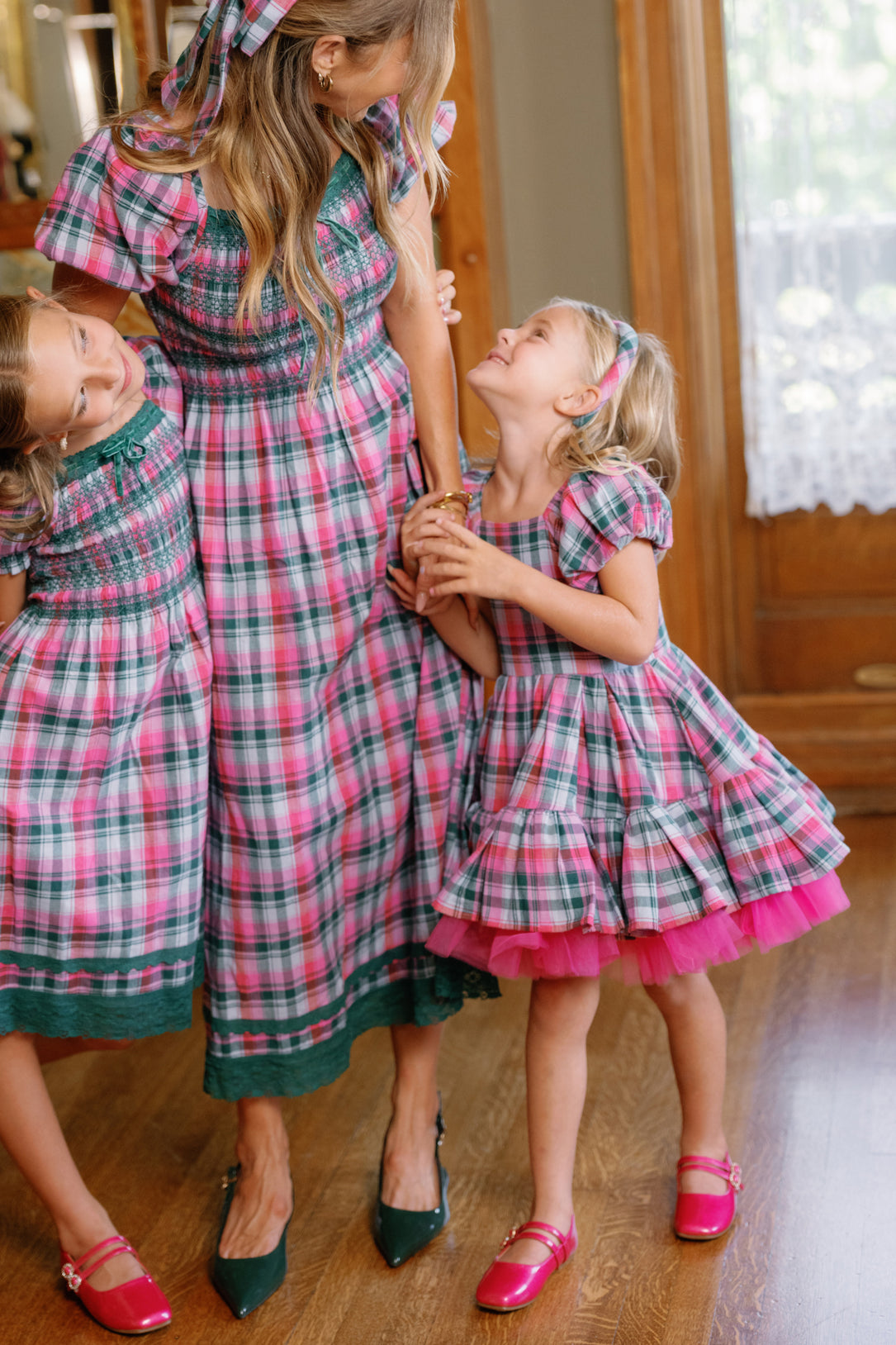 A woman and two young girls wear matching Ivy City Co’s Mini Scottie Dress in Vibrant Plaid—an ideal choice for a girls Christmas dress or holiday party outfit. The trio holds hands and smiles at one another, standing on a wooden floor in a cozy, sunlit room.