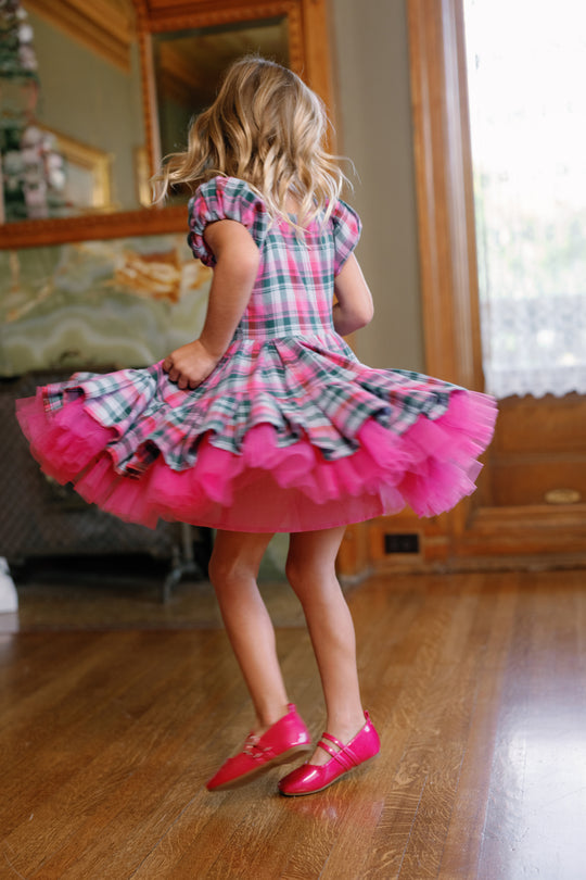 A young girl twirls indoors in the Mini Scottie Dress in Vibrant Plaid by Ivy City Co, her skirt swirling as she stands in pink shoes on a polished wooden floor near a window draped with sheer curtains.