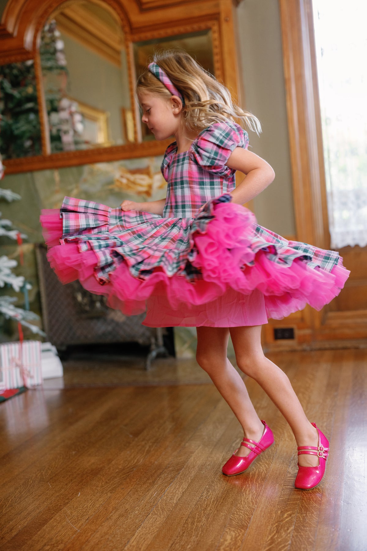 A young girl twirls joyfully in the Ivy City Co Mini Scottie Dress in Vibrant Plaid—a charming holiday dress with a playful pink frilly skirt and matching shoes—in a festively decorated room with wood floors and a sparkling Christmas tree.