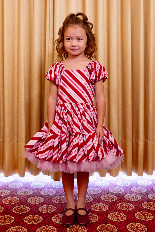 Young girl wearing a red and white striped dress with a ruffled skirt against a beige curtain background.